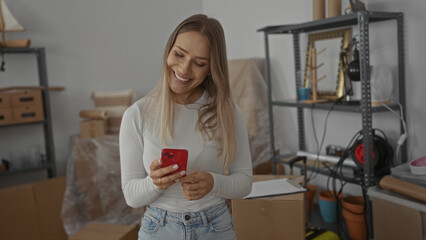 Young woman using smartphone in new home surrounded by unpacked boxes in a living room setting with casual interior elements and personal belongings visible around.