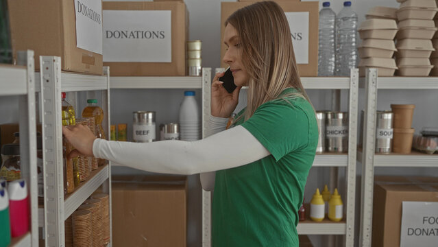 Woman talking on phone inside charity donation center surrounded by shelves filled with various food items and boxes labeled donations indicating volunteer work setting.