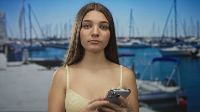 Girl stands on sunlit port pier using smartphone to scroll messages near moored sailboats under clear sky; focus.