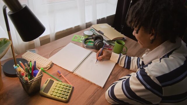 High angle view of creative gen Z teenage black boy making sketch in copybook while sitting at home table with homework