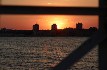 Sunset at sea with silhouettes of residential buildings on the horizon, creating a tranquil...