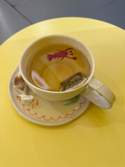 A cup of herbal tea with a tea bag sits on a yellow table, decorated with a whimsical red lobster illustration inside the cup