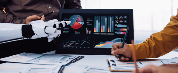 A dynamic office scene shows professionals collaborating with advanced technology. A robotic arm points at detailed graphs, demonstrating innovative business insights and analysis.Noogenesis