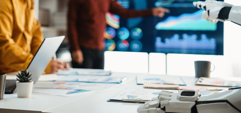 A business meeting in a modern office setting showcases a robotic hand aiding in data analysis. Team members explore charts and graphs, reflecting innovation and collaboration.Noogenesis - Powered by Adobe