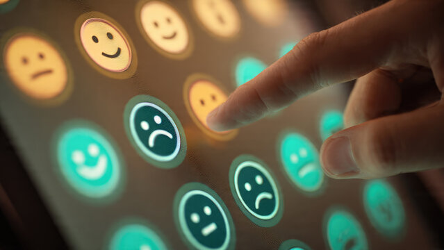 A finger interacts with a digital feedback screen, selecting a neutral emotion from an array of happy, neutral, and sad emoticons to provide user sentiment. - Powered by Adobe