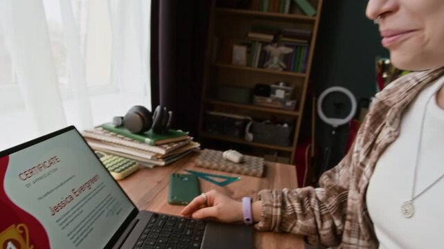 Handheld shot of young gen Z Caucasian girl being excited while sitting in front of laptop screen getting certificate of appreciation at home desk