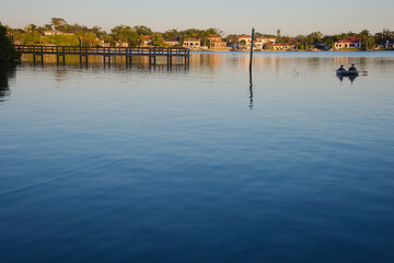 Two Kayakers Paddle Calm River by Docks at Sunset, Reflective Water and Lush Greenery. Coffee Pot Bayou St. Petersburg, FL. Glide across a calm river beside a wooden dock, with green trees and suburba