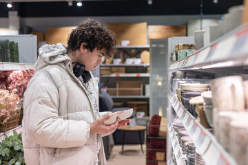 Independent Young Adult Man Shopping for First Home Essentials and Kitchenware. Modern Lifestyle, Separation from Parents, and Moving Concept. Customer Choosing Dishes at a Furniture Store