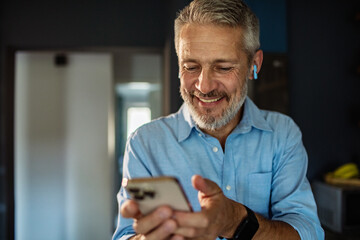 Mature man smiling at smartphone with earbuds at home