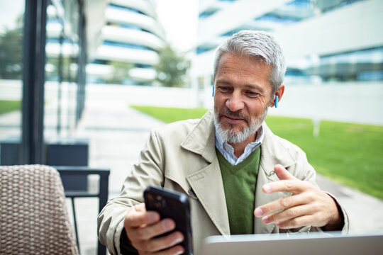 Mature man video calling on smartphone at outdoor office, smiling
