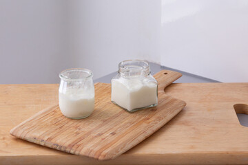 Two small glass jars filled with thick homemade yogurt stand on a wooden cutting board, illuminated...
