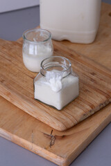 A square glass jar filled with thick homemade yogurt sits on a wooden board, with a small portion gently dripping over the edge. A second glass jar and a large milk container are visible 