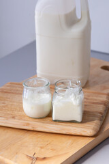 Two glass jars filled with fresh homemade yogurt or kefir sit on a wooden cutting board, with a large milk bottle behind them. The bright natural lighting highlights the creamy texture and purity 