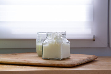 A square glass jar filled with thick homemade yogurt stands in front of a second jar with a more liquid consistency. Soft window light brightens the scene, highlighting the yogurt’s natural texture.