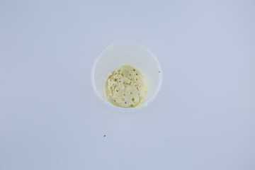 A small portion of spoiled fermented dairy sits at the bottom of a transparent plastic cup, showing visible mold spots, curdled texture, and early decomposition on a sterile white background