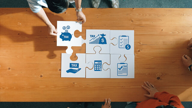 Diverse group collaborates on tax strategy as they arrange puzzle pieces with financial icons on a wooden table, illustrating concepts of planning and budgeting for better financial outcomes. Amity