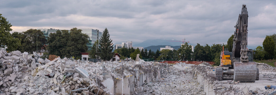 A demolition site filled with concrete rubble and twisted rebar. A large excavator stands ready amidst the debris, framed by urban office buildings and distant mountains under a cloudy sky in i ka.