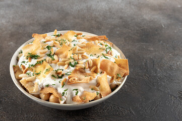 Fatteh with yogurt, crispy pita bread, chickpeas, pine nuts, and herbs in a bowl on a gray background. Lebanese cuisine.