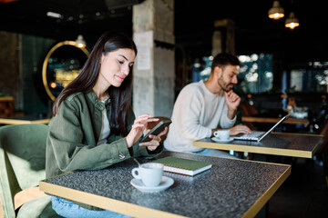 Man and woman smiling, holding coffee while working on a laptop and using a mobile phone  in a cafe