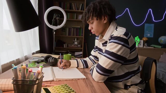Side view of gen Z black teenager drawing sketch with pencil in copybook while sitting at home table in room with neon decor
