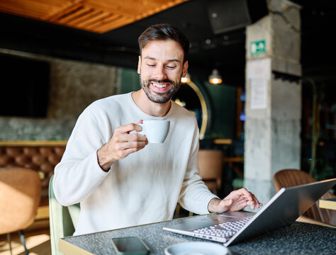 Man smiling, holding coffee while working on a laptop in a cafe