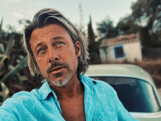 Tanned middle aged man with half long blond hair in blue shirt by classic car at white spanish house in the countryside. Golden hour in Malaga, Andalusia, Spain.