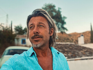 Tanned middle aged man with half long blond hair in blue shirt by classic car at white spanish house in the countryside. Golden hour in Malaga, Andalusia, Spain.