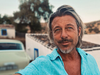 Tanned middle aged man with half long blond hair in blue shirt by classic car at white spanish house in the countryside. Golden hour in Malaga, Andalusia, Spain.