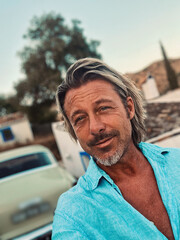 Tanned middle aged man with half long blond hair in blue shirt by classic car at white spanish house in the countryside. Golden hour in Malaga, Andalusia, Spain.