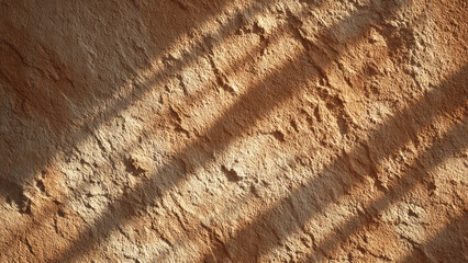 Textured earthy wall with rough surface, illuminated by strong diagonal sunlight creating contrasting shadows and highlighting its uneven contours.