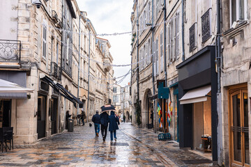 Obraz premium A charming narrow street in La Rochelle featuring historic limestone facades and traditional arcades. An artisan bakery and cafe tables line the wet cobblestone pavement under an overcast sky.