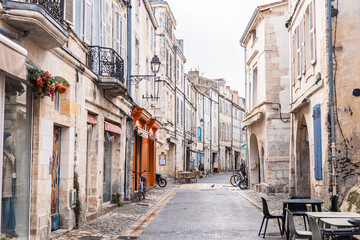 A charming narrow street in La Rochelle featuring historic limestone facades and traditional arcades. An artisan bakery and cafe tables line the wet cobblestone pavement under an overcast sky.