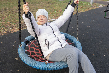 A woman is swinging on a round baby swing on a playground. She is wearing a white jacket and a knitted hat.