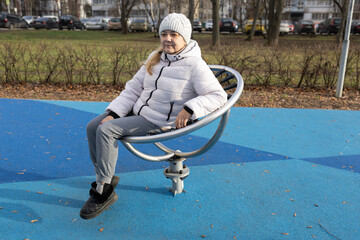 A woman is swinging on a round baby swing on a playground. She is wearing a white jacket and a...