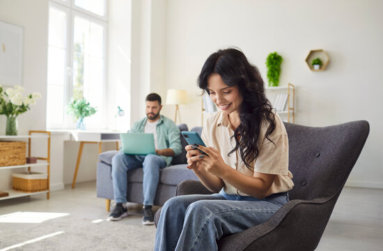 Couple at home using smartphone and laptop. She smiles at messages while he works, highlighting technology and communication shaping relationship distance. Concept tech distracts pair at home.