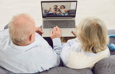 Gray-haired senior couple sitting on sofa in living room at home and having online video call with adult children and granddaughter using laptop. Retirement and communication technology concept.