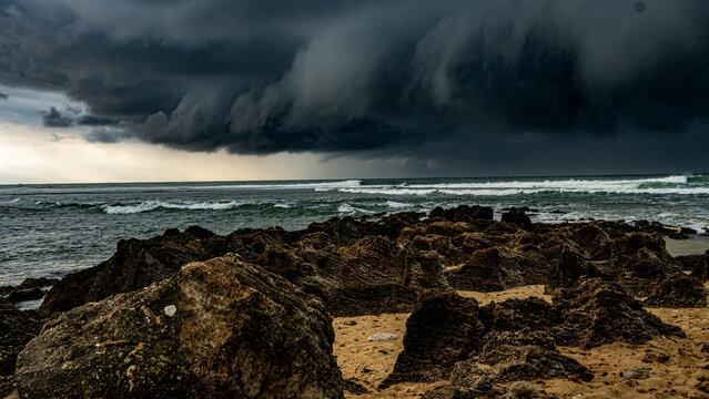 Dark storm clouds over a choppy ocean with jagged rocks and sandy shoreline. Dramatic weather and natural intensity captured in one frame - Powered by Adobe