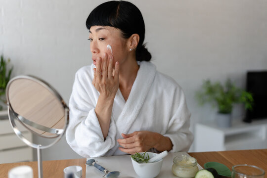 Smiling Asian woman in a white bathrobe applying face cream while looking into a mirror, surrounded by natural skincare products in a relaxed home setting - Powered by Adobe