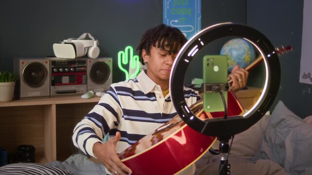 Medium long shot of young African American boy streaming on smartphone in ring light while playing acoustic guitar in cozy bedroom