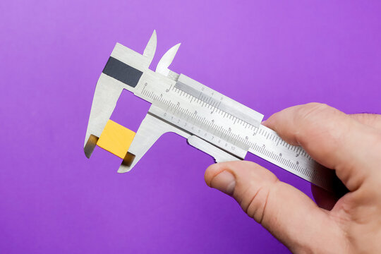 A hand holds a caliper measuring yellow cube violet background. Ruler for precision measuring, functionality in dimensional analysis. Bold contrast to metallic finish and bright yellow cube.
