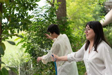 Fototapeta premium Two LGBTQ friends, one with long hair and the other with short hair, enjoy watering their garden in the morning. They share a joyful moment while caring for their plants in the lush outdoor space.