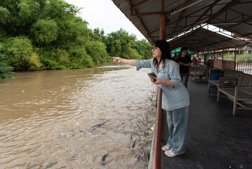Two LGBTQ close friends feeding fish at the alms zone by the river at a Thai temple. The serene activity represents kindness, spiritual connection, and a moment of reflection by the water.