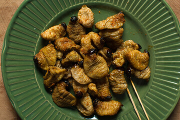 Overhead view of roasted sago worms on a green plate, top view of palm worms on wood background, roasted grub worms in a green dish
