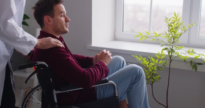 Nurse pushing young disabled male patient in wheelchair in medical clinic. Handicapped man with disability looking at the window in rehabilitation center. Medicine concept. 4k video.