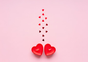 Minimalist flat lay of two unlit red heart-shaped candles with small heart confetti streaming above them on a soft pink background.