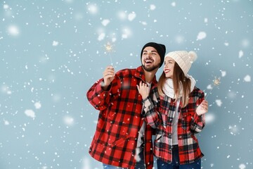 Happy young couple with Christmas sparklers on light blue background