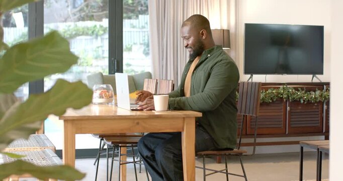 African American mid adult male entering frame sitting at home working on laptop sipping coffee mug