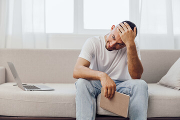 Fototapeta premium Young man sitting on a sofa with a notebook, looking pensive and stressed, surrounded by soft natural light. Ideal for showcasing modern life challenges and mental health themes.