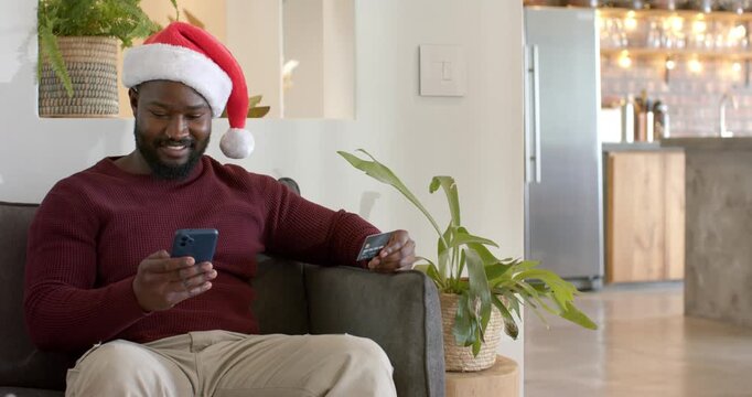 African American man with Santa hat scrolling smartphone, verifying card and paying on sofa