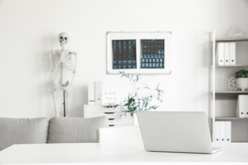 Interior of light hospital with human skeleton model, x-ray images and modern laptop on table